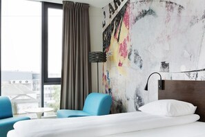 A hotel room with a bed, a blue armchair, a small table, and a view of buildings through a window.