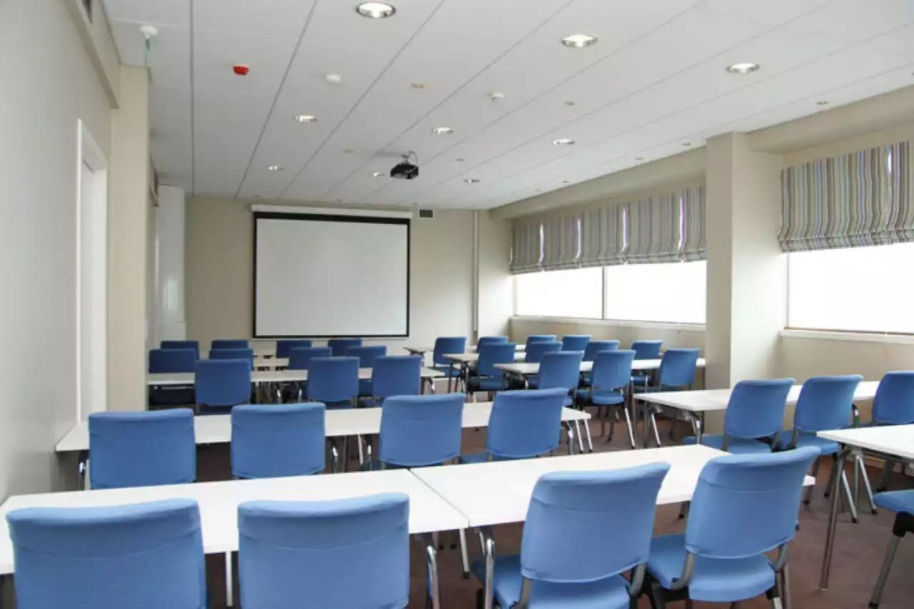 Une salle de conférence avec des rangées de chaises bleues, des tables blanches, un écran de projection et de grandes fenêtres ornées de rideaux à rayures.