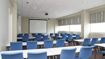 Une salle de conférence avec des rangées de chaises bleues, des tables blanches, un écran de projection et de grandes fenêtres ornées de rideaux à rayures.