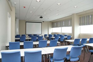 Un salón de conferencias con filas de mesas y sillas, una pantalla de proyector y ventanas con persianas.