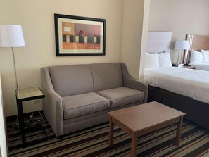 A hotel room with a sofa, a small table, a bed with white bedding, a framed picture on the wall, and a floor lamp. - Best Western Windsor Inn & Suites (Danville)