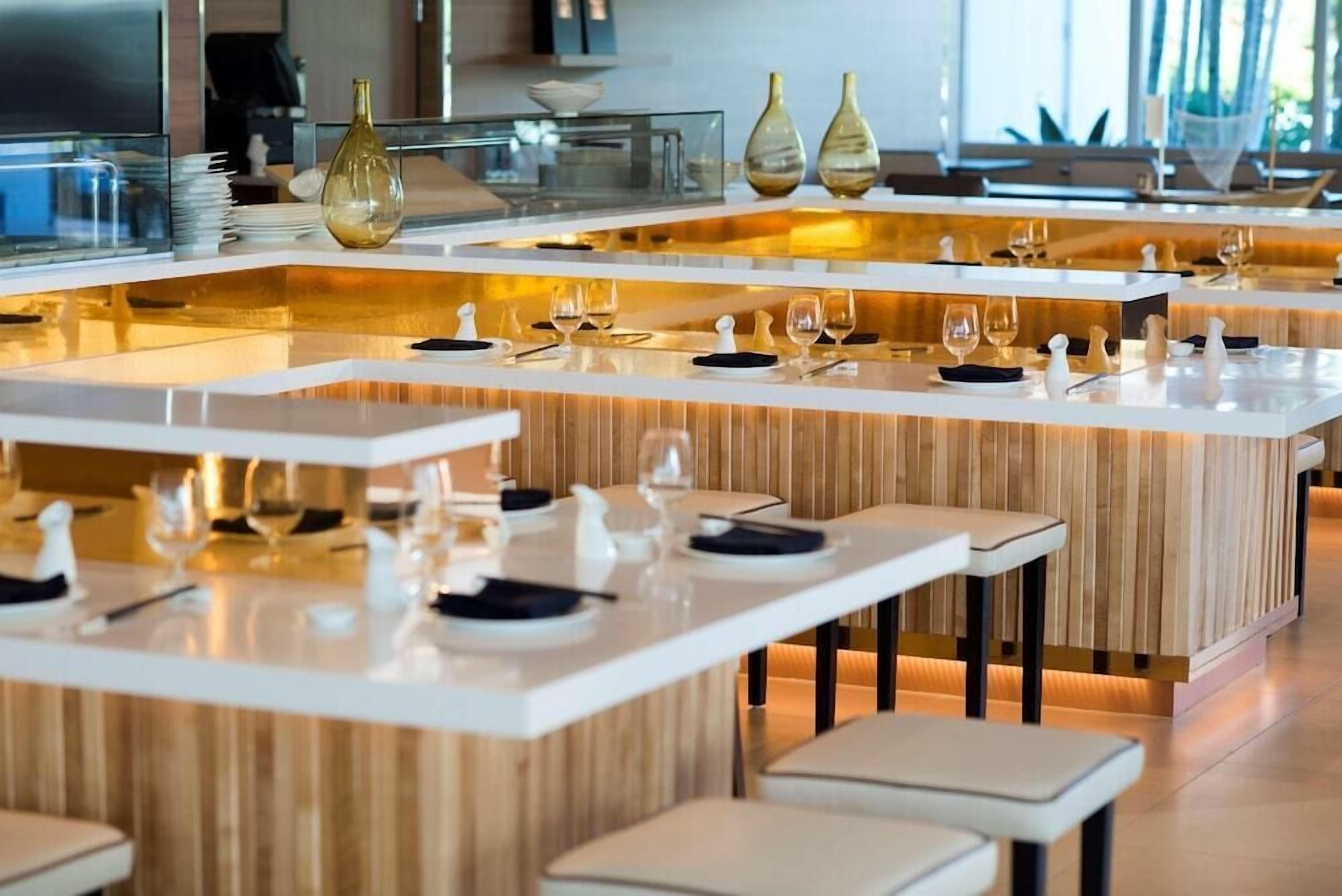 A modern restaurant with multiple dining areas, each with a white table and chairs, and a central bar area with a wooden counter.