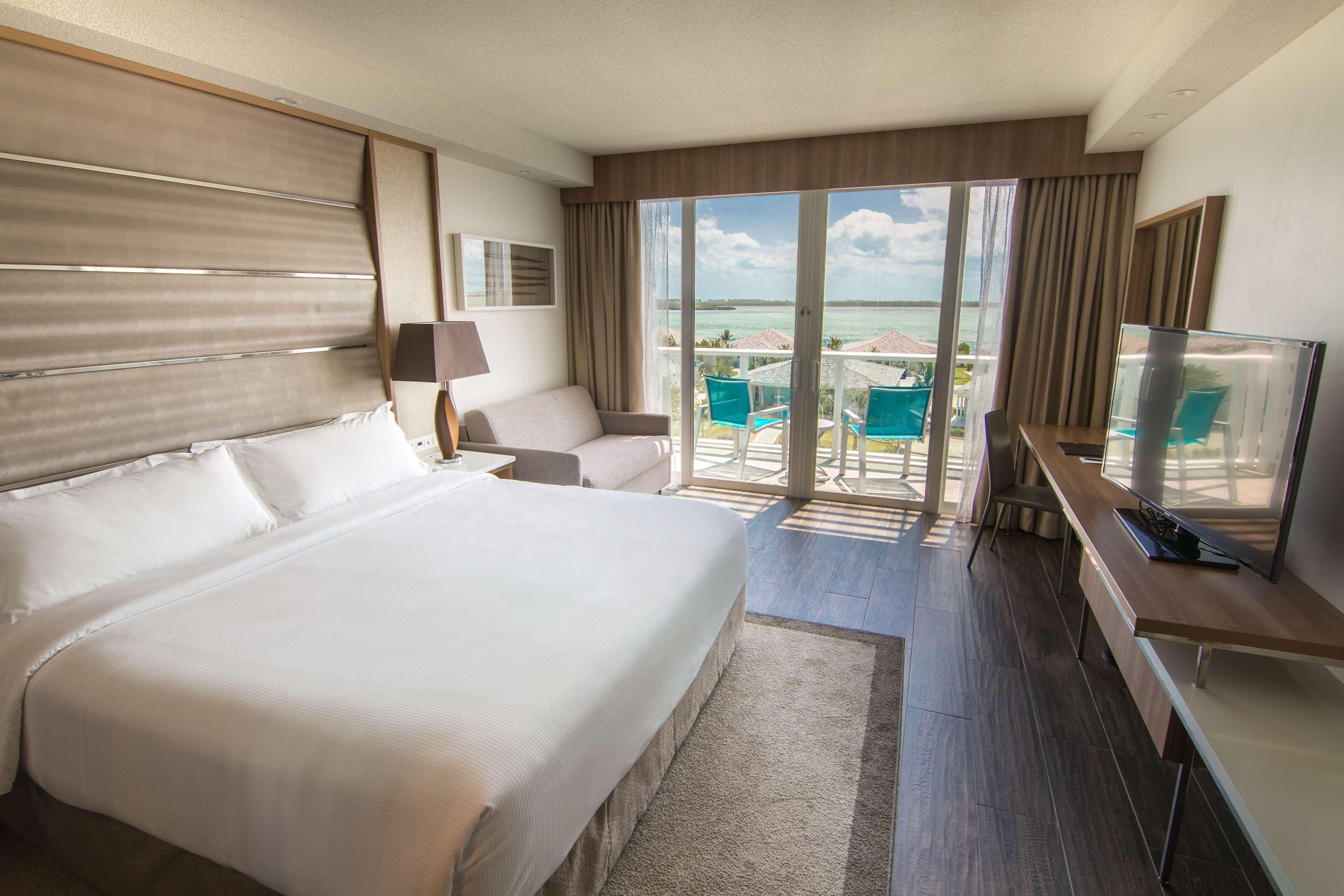Een hotelkamer met een groot bed, een bureau, een stoel, een televisie en een balkon met uitzicht op zee.