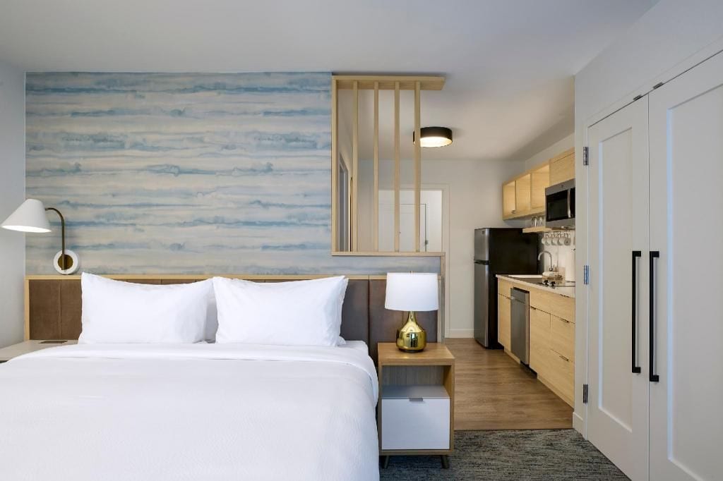 A hotel room with a bed, bedside lamp, a view of the kitchen, and a wall with a blue abstract design.