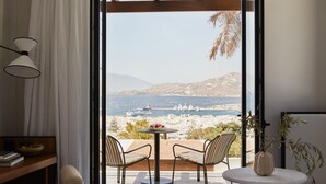 A bedroom with a large bed, a desk, a chair, a small table, and a view of a balcony with chairs and a table. - Anandes Hotel - Adults Only (Mykonos)