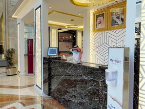 A hotel reception area with a marble counter, a reception desk, and framed pictures on the wall. - Corp Al Khobar Corniche Hotel (Al Khobar)