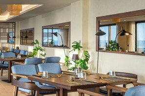 Ein moderner Essbereich mit Holztischen und blauen Stühlen, großen Fenstern und Topfpflanzen.