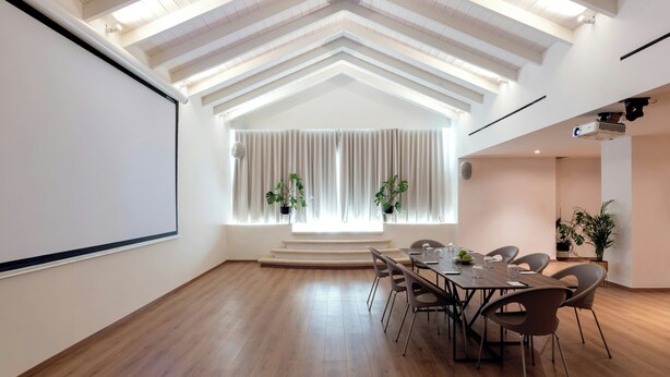 A conference room with a large screen, wooden flooring, a high ceiling with exposed beams, a window with curtains, and several chairs around a table. - Aulus Chania Resort, Curio Collection by Hilton (Chania)