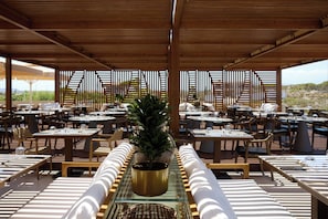 An outdoor dining area with multiple tables and chairs, a central potted plant, and a wooden ceiling structure. - Aulus Chania Resort, Curio Collection by Hilton (Chania)