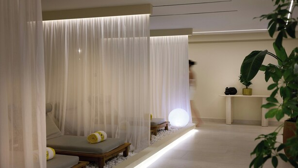 A spa room with two wooden benches, a white curtain, a small table with a plant, and a white floor. - Aulus Chania Resort, Curio Collection by Hilton (Chania)