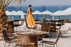 A dining area with wicker chairs and tables, a woman in a dress, and a view of the ocean. - Aulus Chania Resort, Curio Collection by Hilton (Chania)