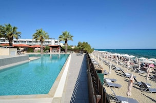 Una piscina con agua cristalina azul, una playa de arena con tumbonas y sombrillas, y un edificio con balcones al fondo.