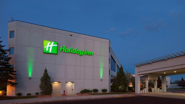 A Holiday Inn hotel with a prominent green logo, a flat facade, and a clear sky. - Wyndham Flint Mi/grand Blanc/bishop Airport/conference Ctr (Flint)