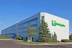 A modern hotel building with a prominent green logo, multiple floors, and a parking lot. - Wyndham Flint Mi/grand Blanc/bishop Airport/conference Ctr (Flint)