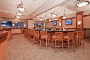 A hotel bar with multiple seating areas, a television screen, and a counter with various bottles. - Wyndham Flint Mi/grand Blanc/bishop Airport/conference Ctr (Flint)