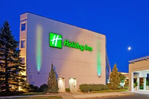 A Holiday Inn hotel building with a prominent green logo, a flat facade, and a well-lit entrance. - Wyndham Flint Mi/grand Blanc/bishop Airport/conference Ctr (Flint)