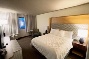 A hotel room with a bed, a desk, a chair, a nightstand, and a window with curtains. - Wyndham Flint Mi/grand Blanc/bishop Airport/conference Ctr (Flint)
