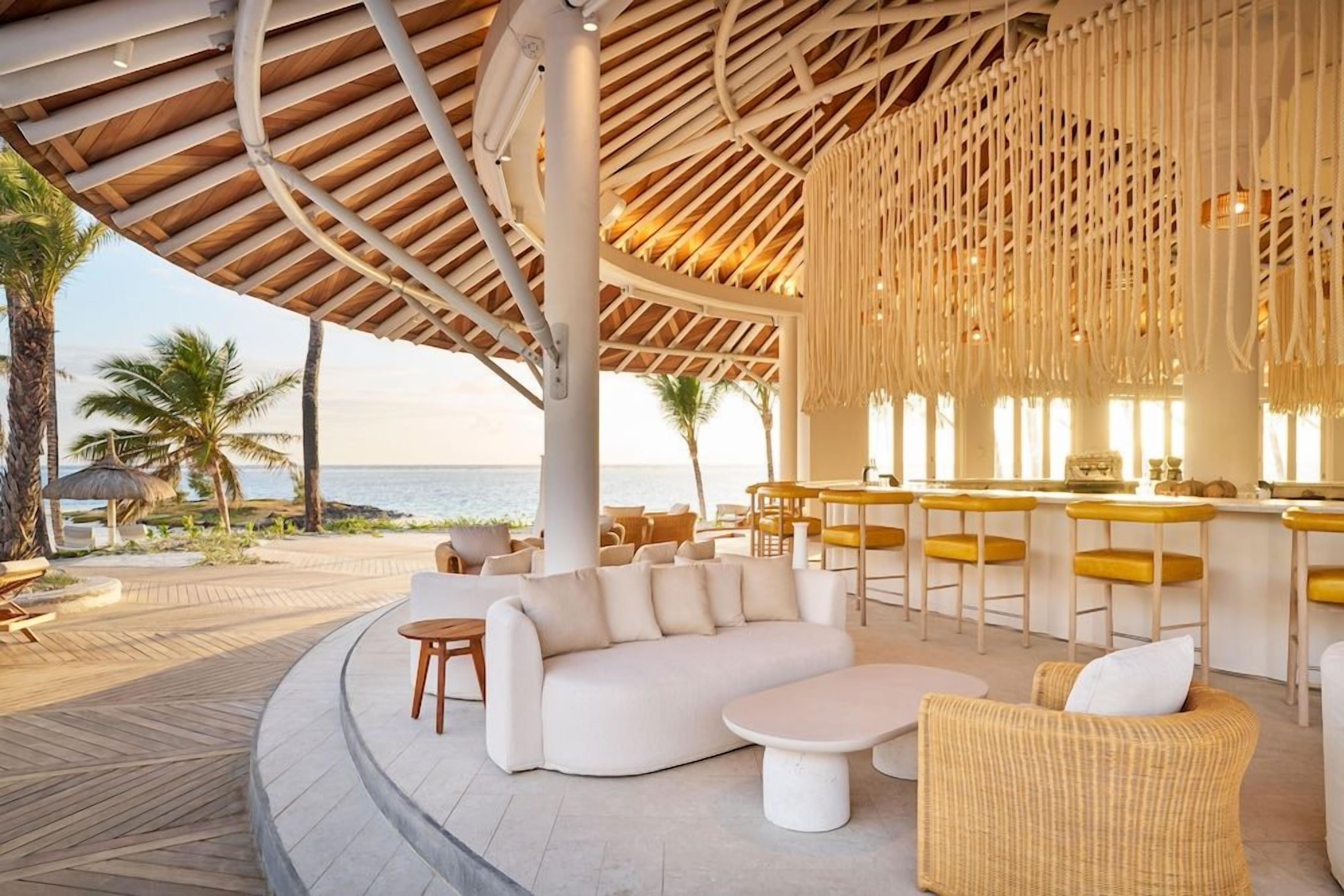 An outdoor lounge area with a curved seating arrangement, a bar with yellow stools, and a view of the sea.