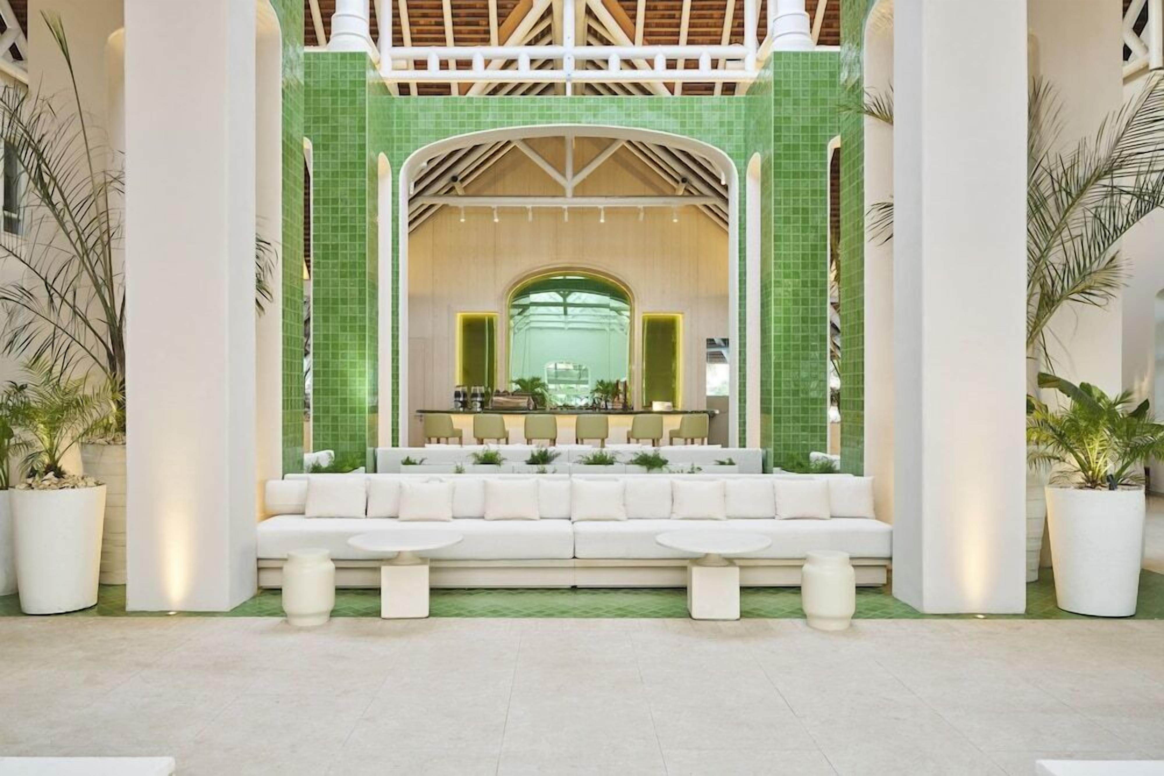 Un vestíbulo con un sofá blanco, dos mesas redondas y una zona de recepción con un muro de azulejos verdes.