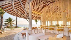 A covered outdoor seating area with a curved sofa, round tables, and a bar with yellow stools. - LUX Belle Mare Resort and Villas (Belle Mare)
