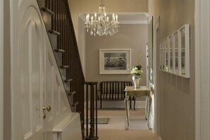 A hallway with a staircase, a chandelier, a framed picture, a bench, and a table with a vase. - Dock House Boutique Hotel (Cape Town)