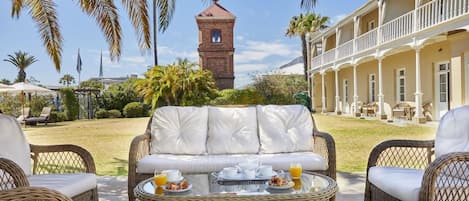 A patio area with wicker furniture, a dining table with breakfast items, and a building with a tower in the background.
