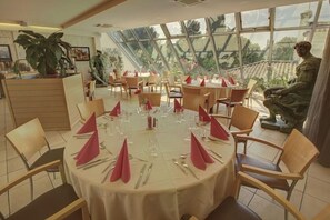 A dining area with round tables set for a meal, surrounded by chairs, with a view of greenery through large windows. - Hotel Atrium (Arles)