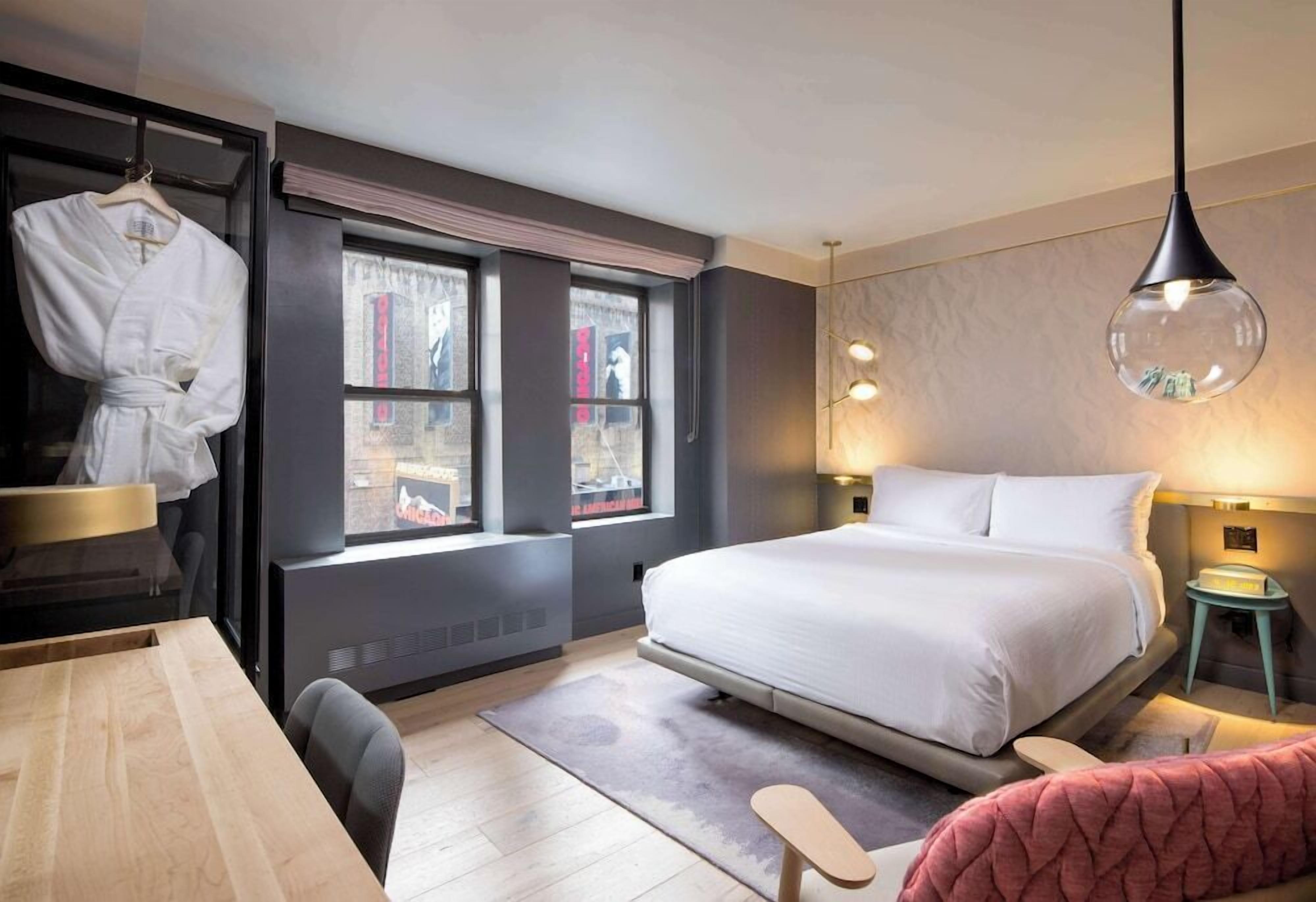 A modern hotel room with a large bed, a desk, a chair, a robe on a hanger, and a window with curtains.