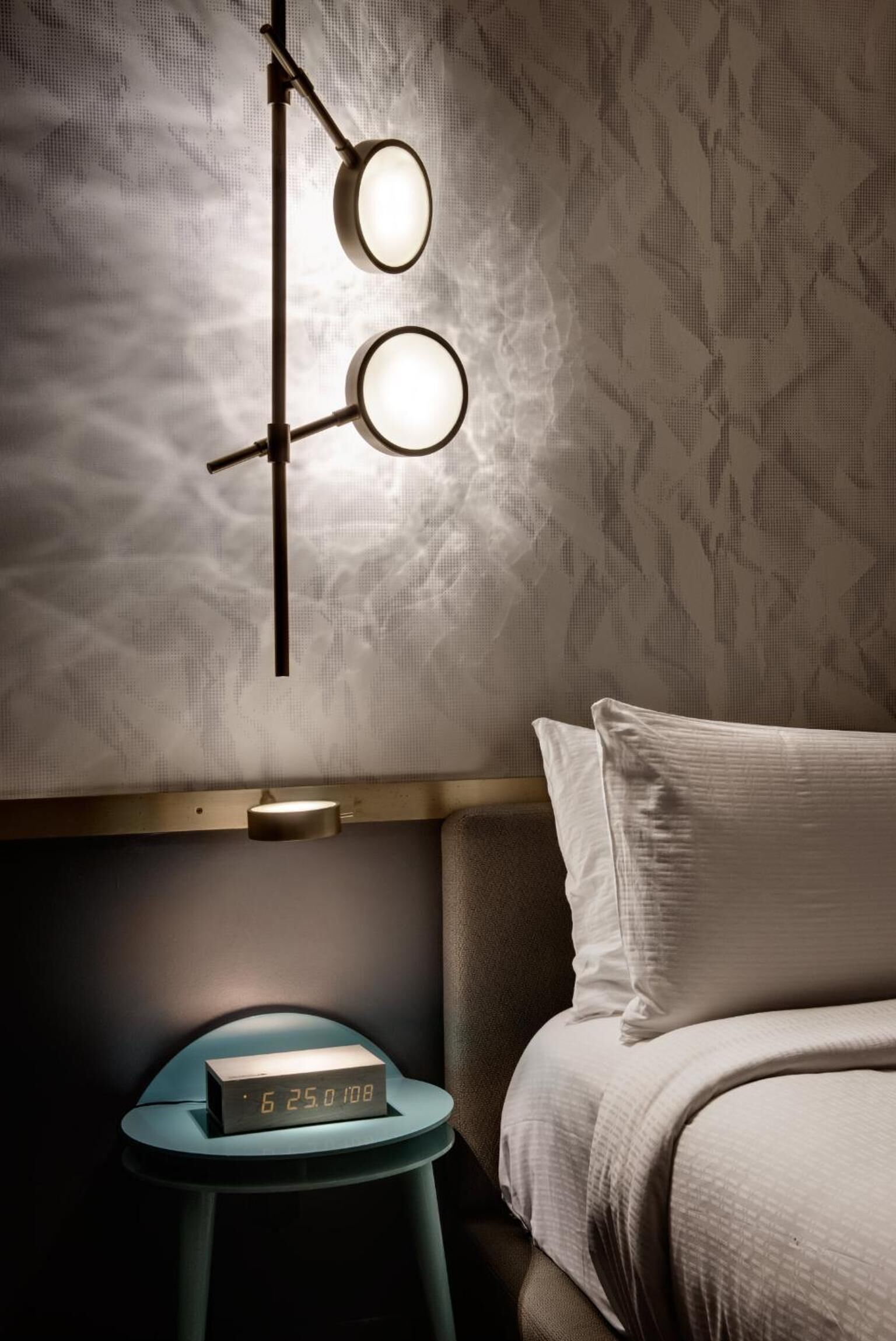 A bedroom with a bed, a bedside table with a digital clock, and a wall-mounted light fixture.