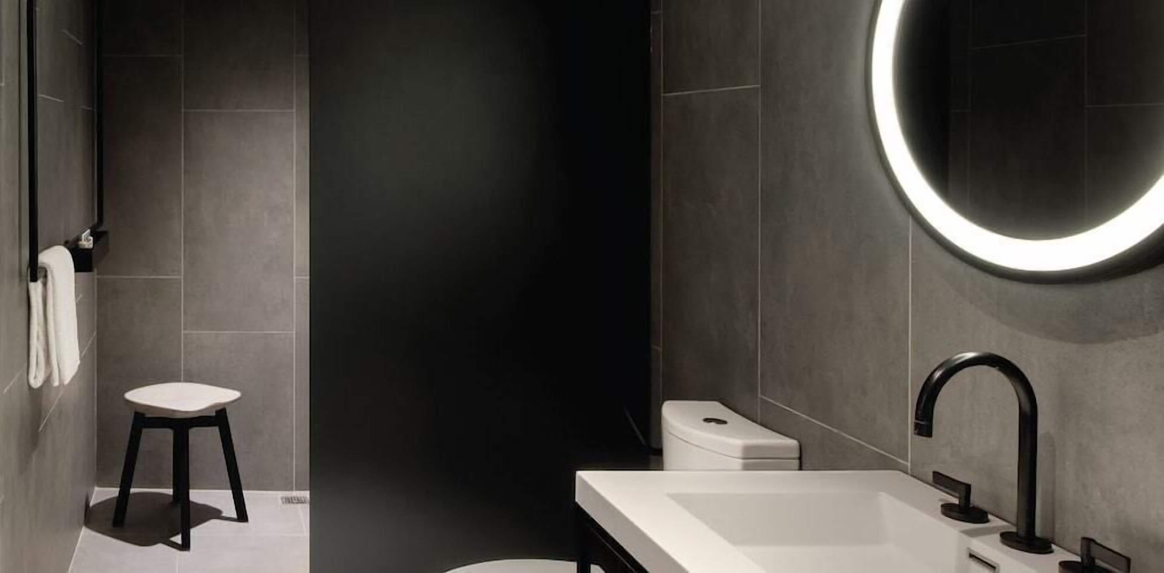 A modern bathroom with a white sink, black faucet, and a round mirror with a light.