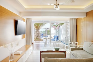 Quarto de hotel moderno com televisão de tela plana, parede de destaque em madeira, mesa de centro de vidro, sofá bege e vista para a área da piscina externa.