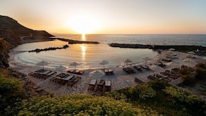 Ein Strandresort mit Liegestühlen und Sonnenschirmen, ein freier Blick auf den Sonnenuntergang über dem Meer und eine felsige Küstenlinie.