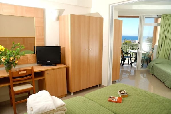 A hotel room with a wooden desk, a television, a green bed, and a balcony with a view of the sea.