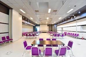 Ein Konferenzraum mit Reihen violetter Stühle, einem zentralen Tisch und einer Projektionsleinwand.
