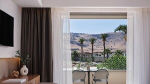 A hotel room with a bed, a desk, a chair, a TV, and a balcony with a view of palm trees and hills.