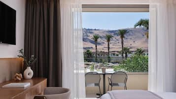 A hotel room with a bed, a desk, a chair, a TV, and a balcony with a view of palm trees and hills.