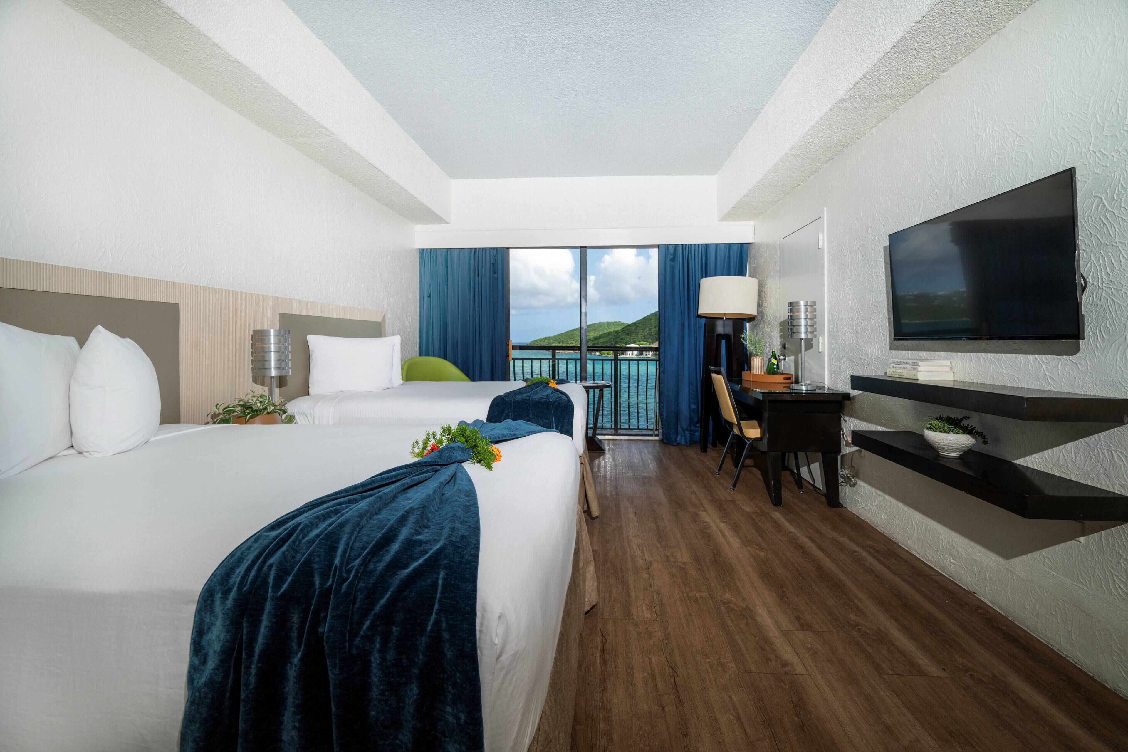 A hotel room with a large bed, a desk, a TV, and a balcony with a view of the sea.