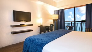 Habitación de hotel con cama, televisión, escritorio, silla, lámpara y balcón con vistas.