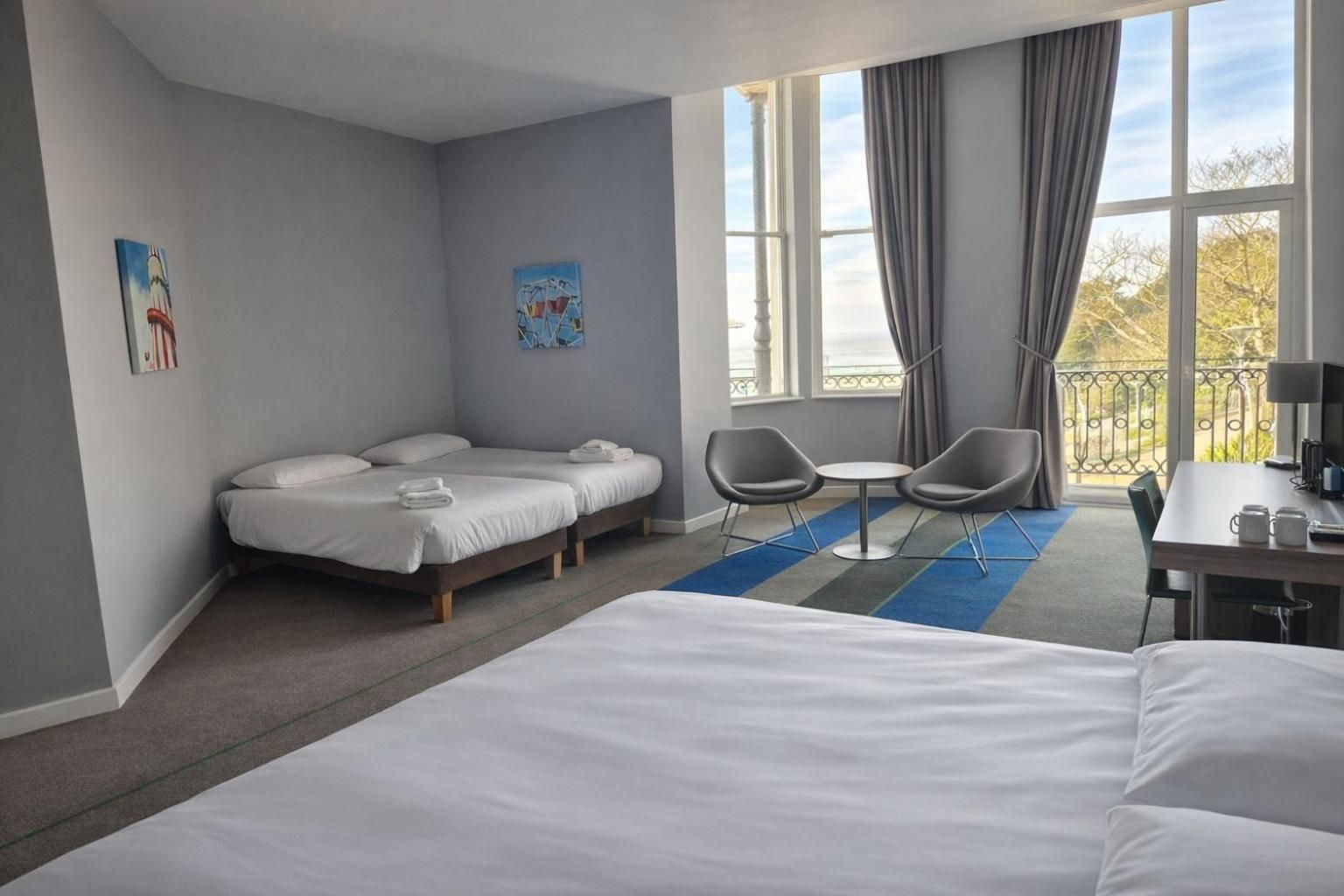A hotel room with a large bed, two smaller beds, a desk with a chair, a small table with two chairs, and a balcony with a railing.