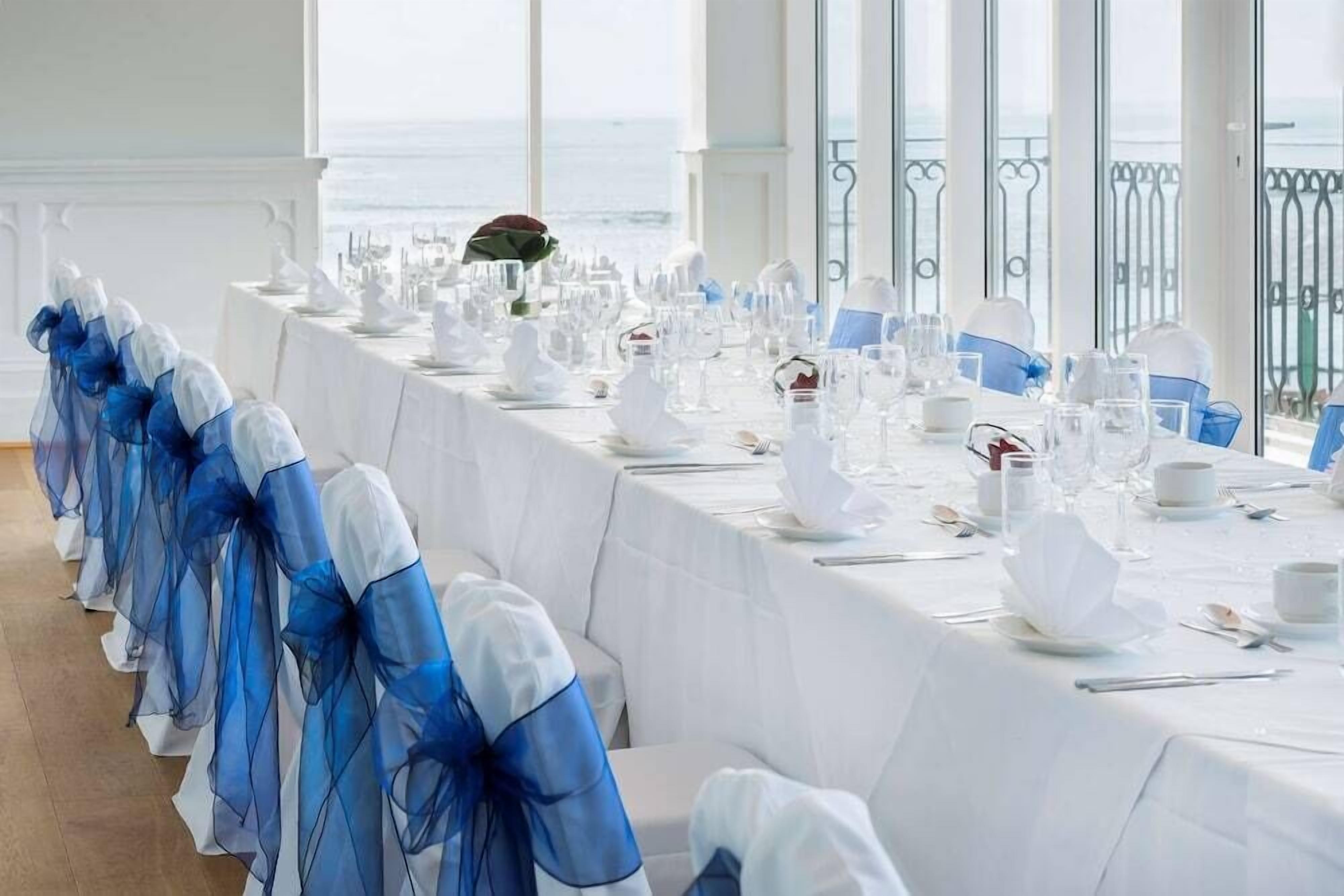 A long banquet table set for a formal event with white tablecloths and blue sashes, overlooking the sea.