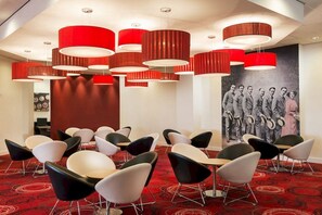 Un salón de conferencias con sillas modernas, lámparas colgantes cilíndricas rojas y una fotografía blanco y negro en la pared.
