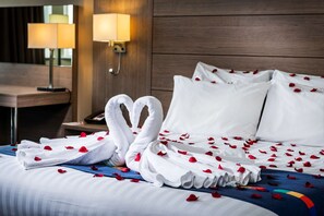 Ett hotellrum med en snyggt bäddad säng, vita kuddar, en handduksdekoration formad som ett hjärta och rosenblad utspridda på sängen.