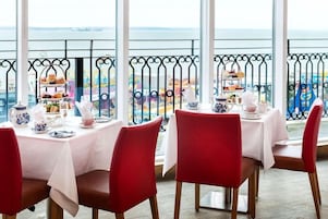 A dining area with tables set for tea service, red chairs, and a view of the sea through large windows.