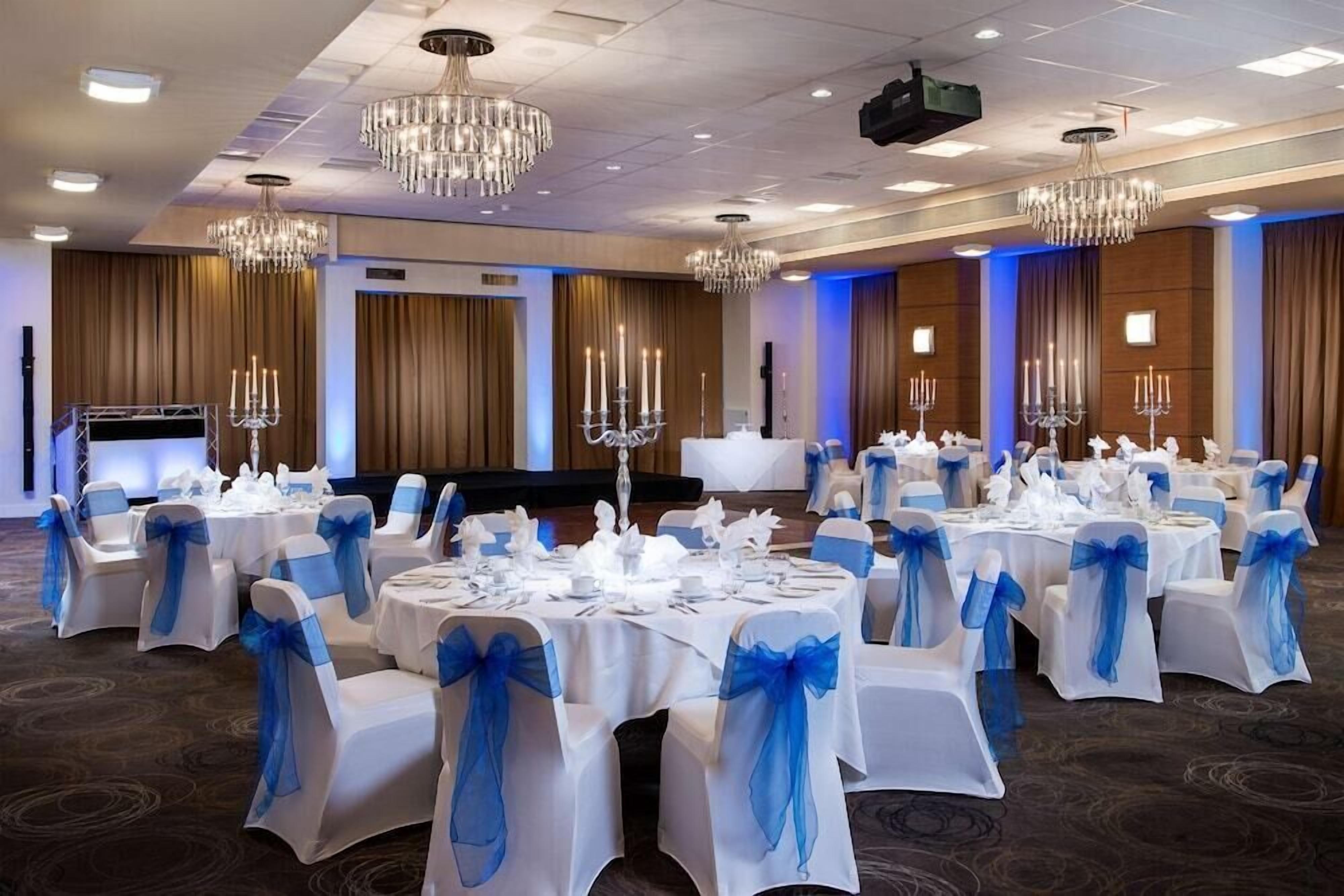 A banquet hall with round tables, blue sashes, chandeliers, and a stage.