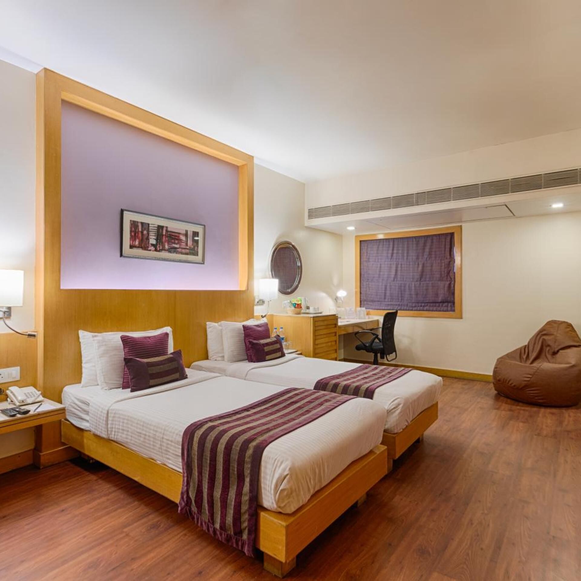 A hotel room with two beds, a desk, a chair, a bean bag, and a wall-mounted picture.