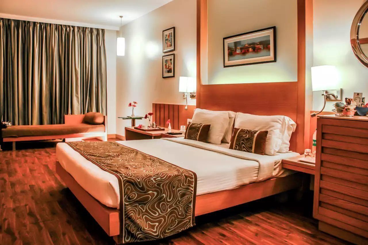 A hotel room with a large bed, a desk, a chair, a nightstand, and a window with curtains.