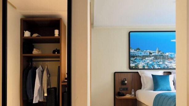 A hotel room with a wardrobe, a bed with a blue pillow, a nightstand, and a painting of a coastal town. - MIA Urban Agadir (Agadir)