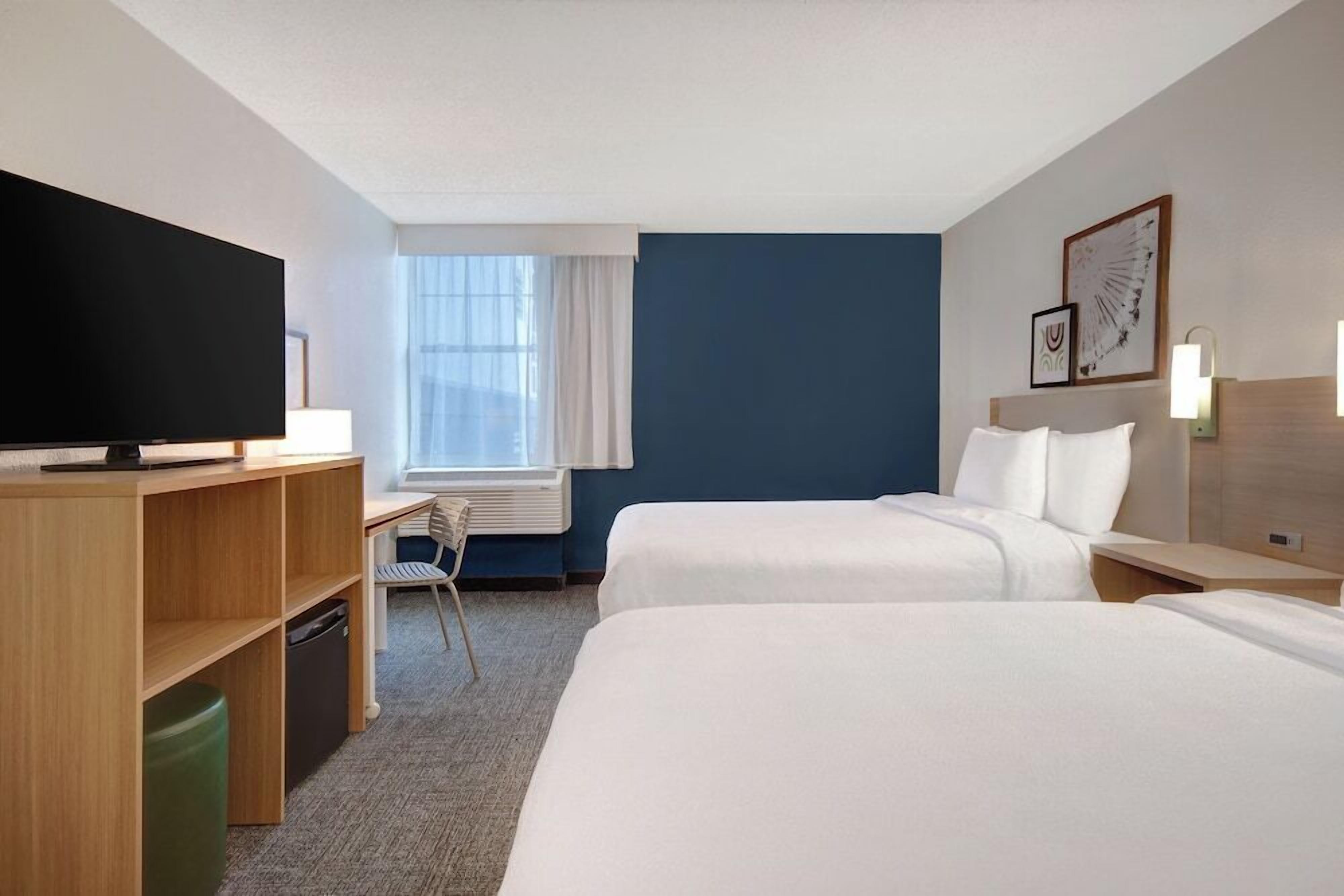 A hotel room with two beds, a desk, a television, and a window with curtains.