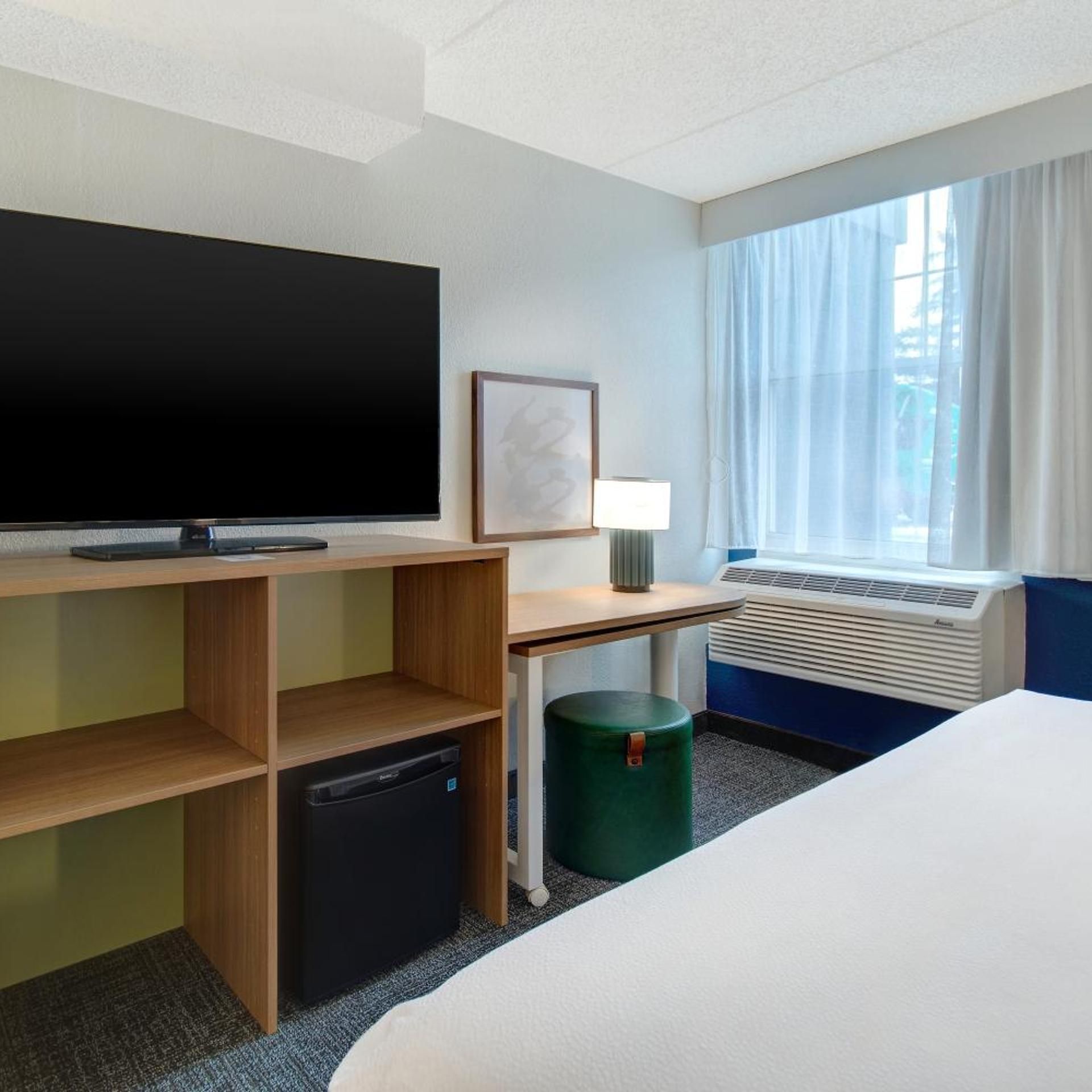 Ein Hotelzimmer mit Flachbildfernseher, Schreibtisch mit Lampe, kleiner Heizung, grünem Mülleimer und einem Bett.