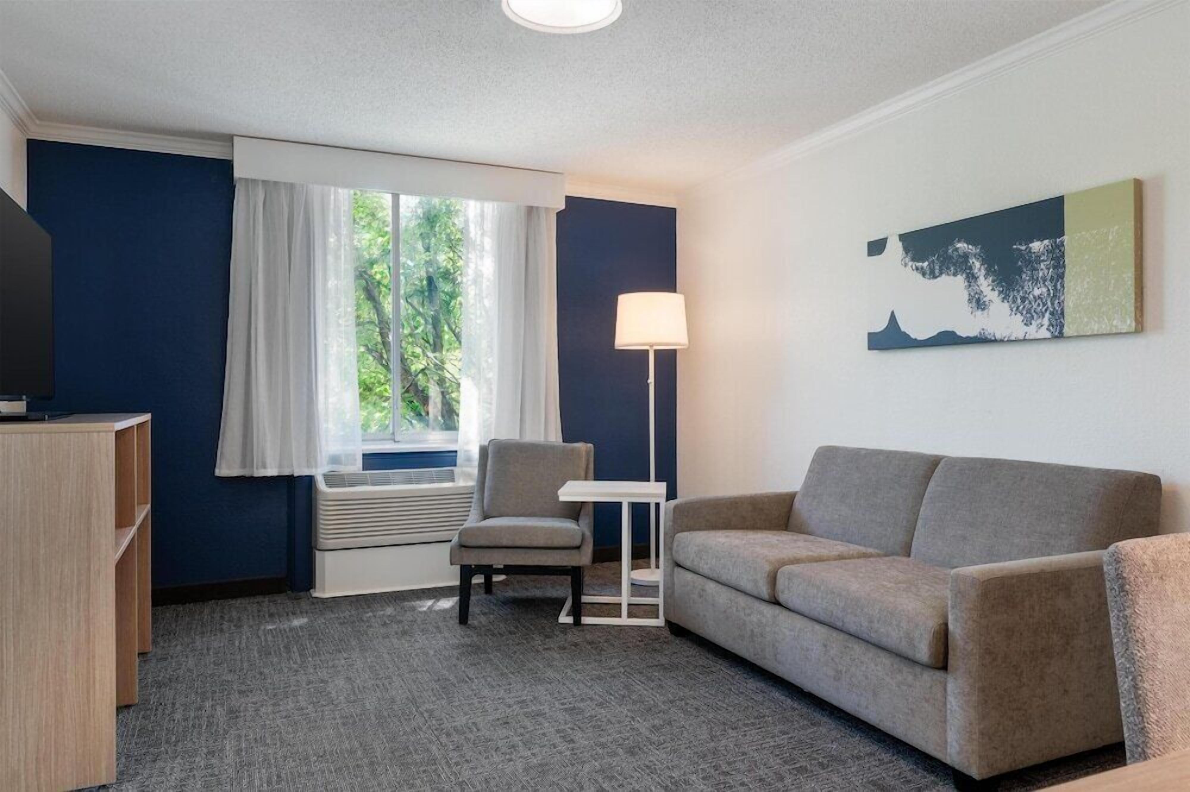 A hotel room with a sofa, a chair, a small table, a lamp, and a window with curtains.
